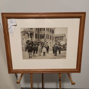 Framed black-and-white vintage photograph of a small-town parade on Orne Street in Marblehead, Massachusetts. The image shows a man on horseback wearing a star badge, several men in early 20th-century attire, an American flag bearer, and a child holding a sign that reads “Ruff for Bill.” The group stands in front of classic New England-style houses, with parked cars visible in the background.